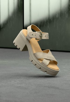 Sandale beige à talon chunky avec larges brides croisées et boucle ajustable, présentée sur un fond vert pâle et gris.