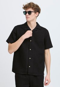 Cămașă neagră cu mânecă scurtă, având un model texturat de tip wafel, cu guler, nasturi bej și o croială lejeră. Poartă ochelari rotunzi de soare.