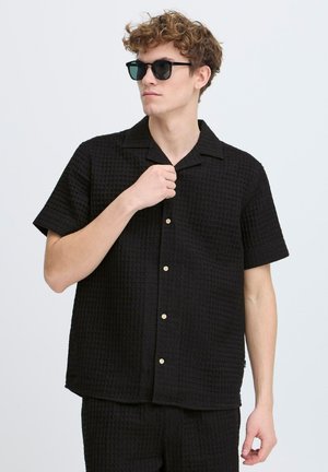 Chemise noire à manches courtes avec un motif en gaufre texturé, dotée d'un col, de boutons beiges et d'une coupe décontractée. Portée avec des lunettes de soleil rondes.