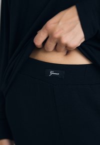 Eine Hand hebt ein schwarzes Hemd an, um den Taillenausschnitt einer schwarzen Hose mit sichtbarem "Guess"-Markenlabel am Bund zu zeigen.