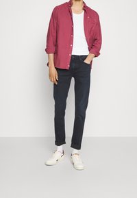 Maroon button-up shirt over a white tank top, paired with dark jeans and white sneakers with red accents. Casual fit and rolled sleeves.