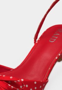 Chaussure slingback rouge à pois blancs, bout pointu et petit talon, montrant la semelle intérieure avec le nom de la marque "RAYD".