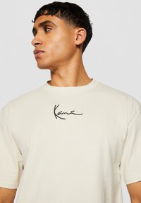 Cream cotton t-shirt featuring a round neck, short sleeves, and a black embroidered signature on the chest. Smooth texture, relaxed fit.