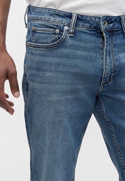Jean en denim bleu moyen porté avec une chemise blanche, montrant le côté avant gauche, les détails de la poche, un bouton et une partie de la main gauche d'une personne.