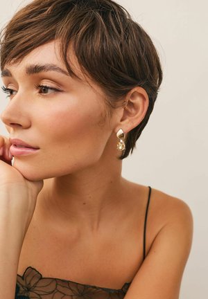 Goldene Ohrringe mit einem Tropfen-Design, das einen klaren Edelstein zeigt. Das Model hat kurze, zerzauste Haare und trägt ein schwarzes, durchsichtiges Oberteil.