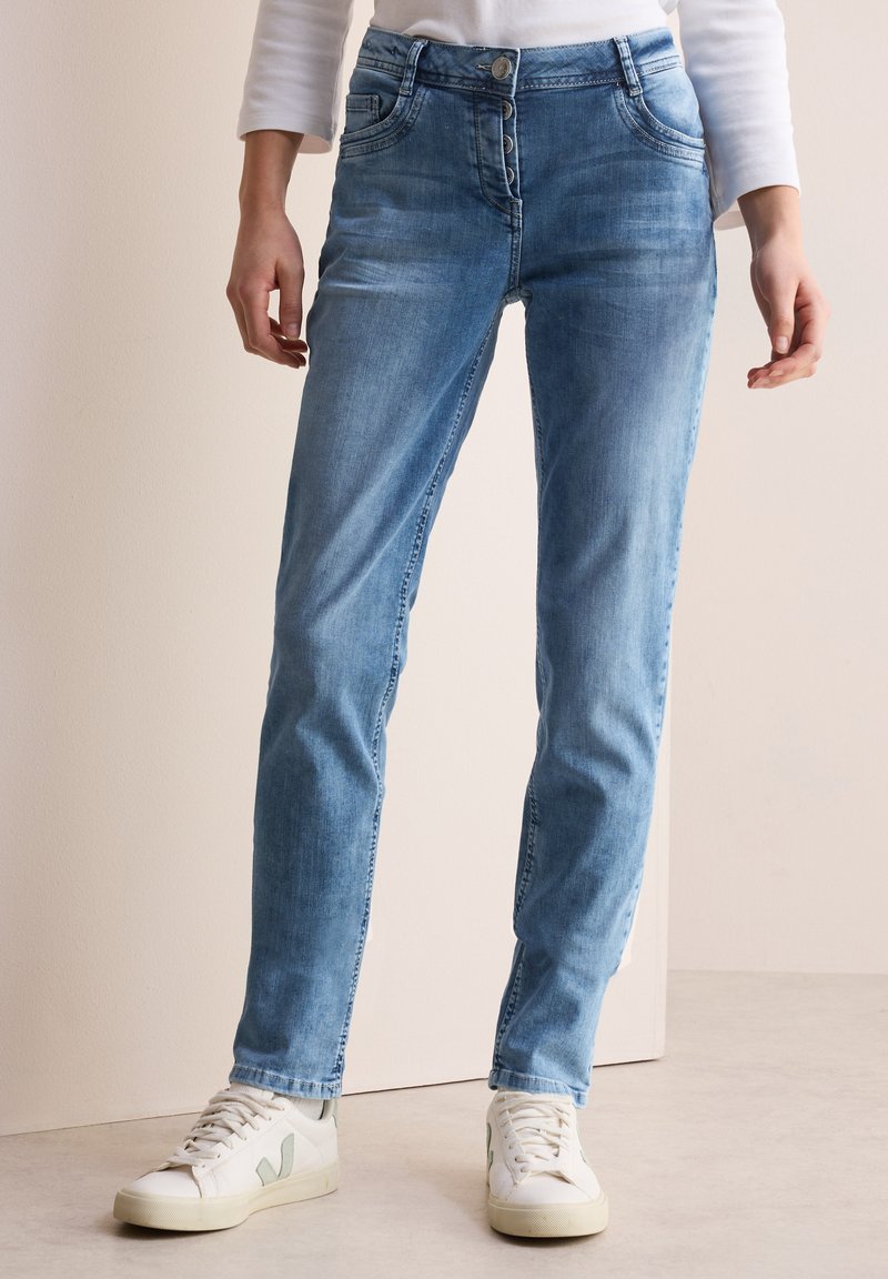 Jeans de mezclilla azul claro con un diseño de pierna recta, que cuentan con una tapeta de botones, bolsillos y un sutil desgaste, combinados con zapatillas blancas.