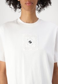 Weiße Baumwoll-T-Shirt mit einem strukturierten Spitzenpatch mit einem schwarzen gestickten Design in der Mitte. Rundhals und kurze Ärmel.