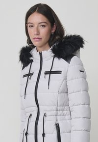 Chaqueta acolchada blanca con capucha de piel sintética negra, bolsillos con cremallera, textura acolchada y cordón ajustable en la cintura y la capucha.