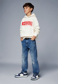Felpa beige con logo "DIESEL" rosso, jeans blu strappati e sneakers blu/bianco. Modello in piedi, mani in tasca, su sfondo grigio.