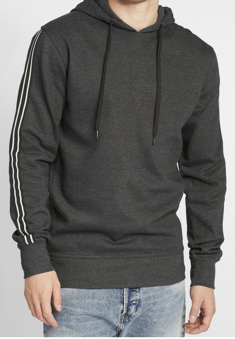 Homme portant un sweat à capuche gris foncé avec des cordons noirs et deux bandes blanches sur chaque manche, associé à un jean bleu clair.