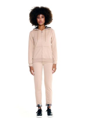 Une femme aux cheveux bouclés pose sur un fond blanc, portant un sweat à capuche beige zippé, un pantalon assorti et des baskets noires.