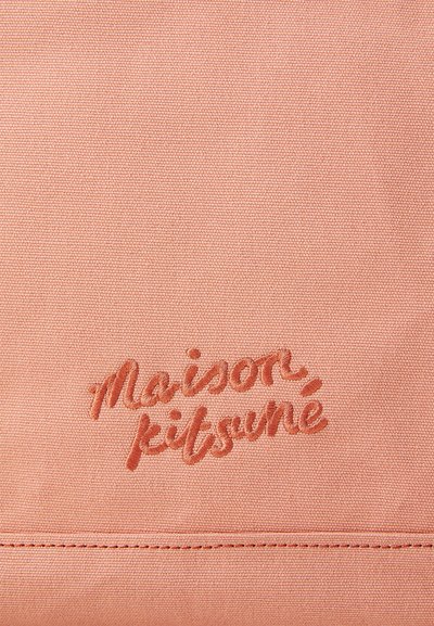 Persiku krāsas audums ar izšūtu "Maison Kitsuné" logo tumši persiku krāsā. Auduma faktūra ir gluda ar redzamu šuvju detaļu.