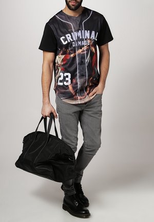 Homme portant un t-shirt graphique noir et gris avec l'inscription « CRIMINAL DAMAGE 23 », un jean gris, des bottes noires, tenant un grand sac de sport noir.
