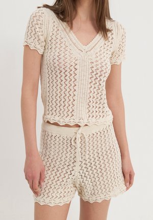 Femme portant un haut à manches courtes en maille de couleur crème et un short taille haute assorti avec cordon de serrage, présentant un motif en crochet ajouré.