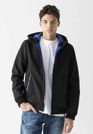 Black softshell jacket with a blue hood interior, zippered pockets, and ribbed cuffs. Layered over a white t-shirt. Smooth texture.