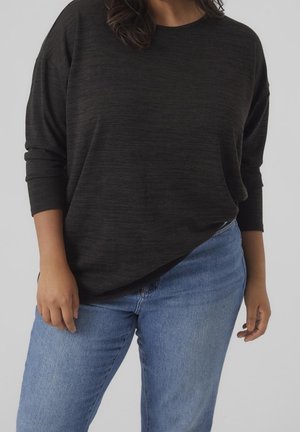 Person iført en løs sort langærmet top og lyseblå denimjeans, stående mod en ensfarvet lys grå baggrund.