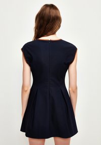 Nainen, jolla on pitkät ruskeat hiukset, pukeutuneena ilman hihoja olevaan tummansiniseen mekkoon, jossa on ruskea reunus, seisoo selin yksinkertaista vaaleaa taustaa vasten.