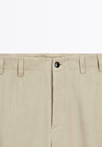 Pantaloni di lino beige con frontale liscio, dotati di due tasche anteriori e un singolo bottone nero in vita per la chiusura.