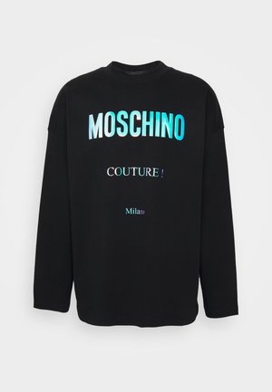 Henkilö, joka pukeutunut mustaan Moschino Couture Milano -merkin pitkähihainen paita, tummiin housuihin ja mustiin lenkkareihin, joissa on valkoiset liekkikuvioinnit.