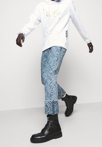 Vit huvtröja med guldtext, urblekta blå denimjeans med logomönster och svarta läderskor med snörning och kraftiga sulor.