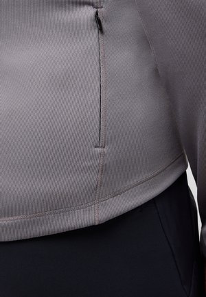 Grijze geribbelde stof top met een ritssluiting aan de zijkant, boven een donkere tailleband, die een strakke en moderne uitstraling benadrukt.