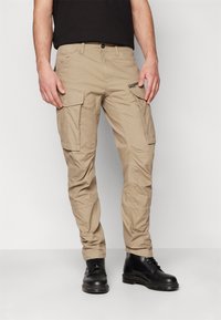 Beige Cargo-Hosen mit mehreren Taschen, schmal zulaufenden Beinen und einer strukturierten Stoffoberfläche. Kombiniert mit schwarzen Schnürstiefeln für einen Kontrast.
