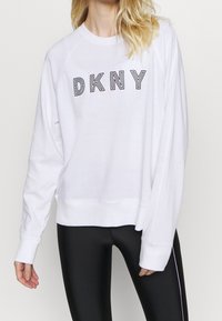 Bílá mikina s černým logem "DKNY", volného střihu, s dlouhými raglánovými rukávy a ribbovanými manžetami a spodním lemem. Vzhledem k černým legínám.