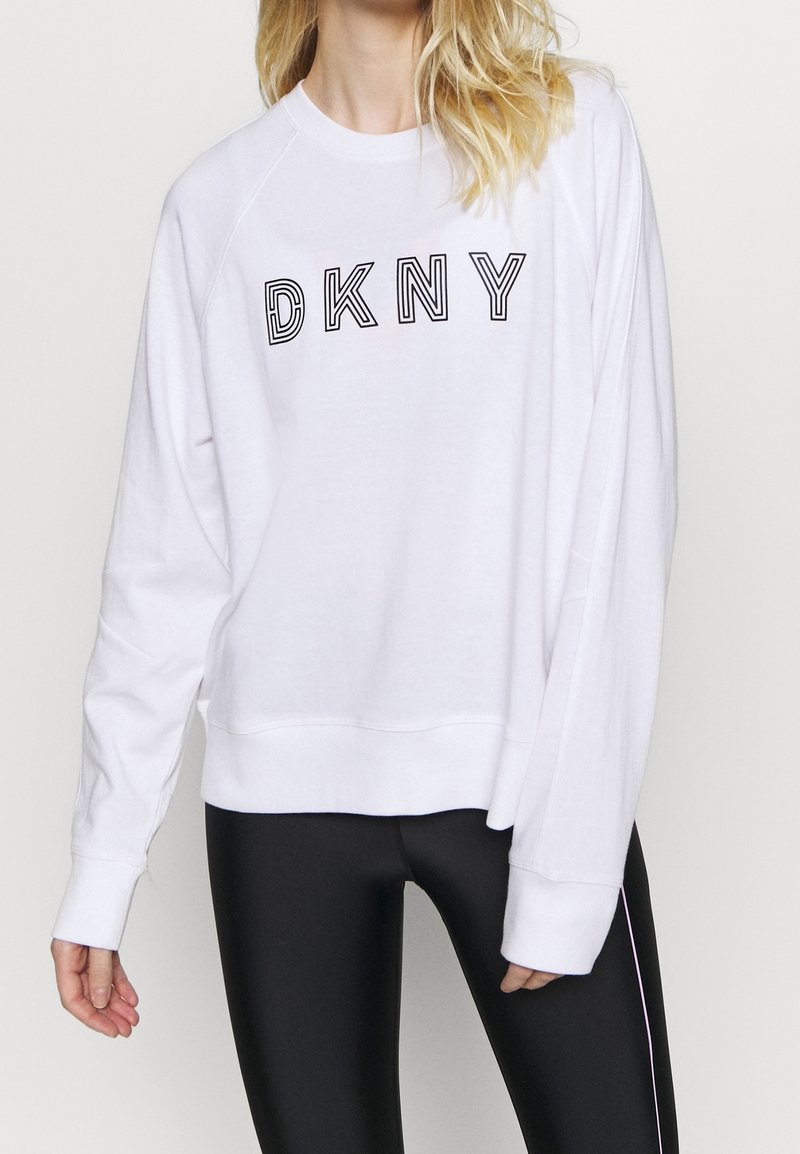 Bílá mikina s černým logem "DKNY", volného střihu, s dlouhými raglánovými rukávy a ribbovanými manžetami a spodním lemem. Vzhledem k černým legínám.