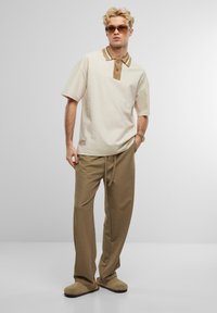 Cremfarbendes Poloshirt mit einem strukturierten braunen Kragen, kombiniert mit lockeren sandfarbenen Hosen und beigefarbenen Slippern. Minimalistisches Design mit einem Logotag.