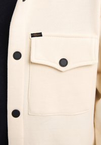 Cárdigan crema con un bolsillo parche y botones negros. Textura de tela suave con una etiqueta oculta en el borde del bolsillo.