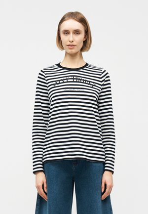 Frau mit kurzem blondem Haar, die ein schwarz-weiß gestreiftes Langarmshirt mit Text und blaue Jeans trägt, stehend vor einem weißen Hintergrund.