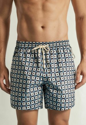 Torace maschile che indossa pantaloncini da bagno decorati con un design geometrico blu e bianco e un laccio bianco in vita.