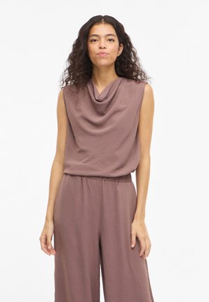 Kvinne med langt krøllete hår iført ermeløs mauve topp med drapert hals og matchende bukser med vid ben, står foran hvit bakgrunn.