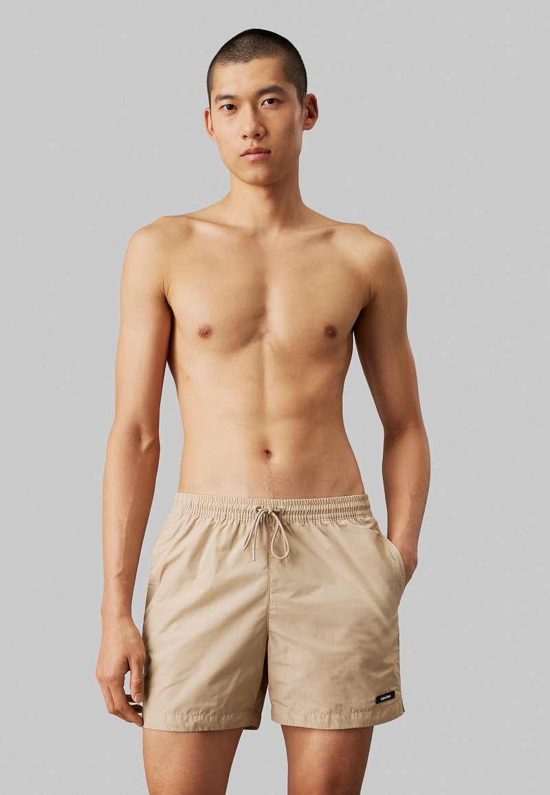 Costume da bagno beige con vita elastica e cordino; presenta due tasche laterali e un piccolo etichetta del marchio nella parte anteriore sinistra in basso.