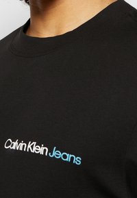 Black crewneck shirt with "Calvin Klein Jeans" logo printed in white and blue on the upper chest, worn by a person with visible neck and jawline.