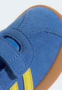 Chaussure en daim bleu avec une sangle à crochets, présentant des rayures jaunes, une semelle en caoutchouc et une surface texturée, mettant en avant des coutures détaillées.