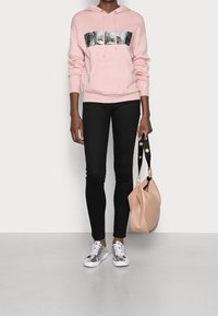Persona che indossa un hoodie rosa con logo GUESS, pantaloni attillati neri, sneakers argentate, mentre tiene una borsa beige con tracolla nera su uno sfondo bianco.