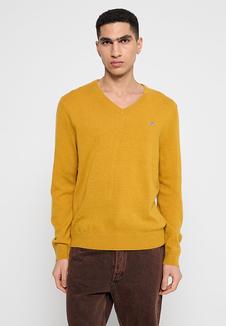 Långärmad senapsgul v-ringad tröja i mjukt stickat material med ribbade mudd och nederkant, med en liten broderad logga på bröstet.