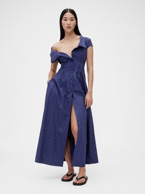 Lang blå kjole med hvite prikker, av-skuldre design, knapplukking foran og sideslitt. Stoffet ser glatt ut med en strukturert silhuett.