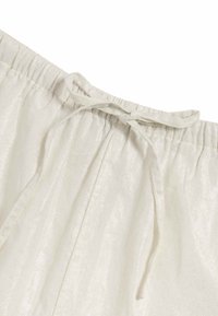 Cintura elastica beige chiaro con dettaglio a lacci. Il tessuto ha una sottile lucentezza e una consistenza morbida, caratterizzato da tessuto arricciato in vita.