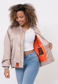 Bomber jacket beige chiaro con zip frontale, polsini a costine e interno arancione. Presenta un'etichetta del marchio all'interno e una texture liscia e lucida.