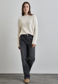 Suéter esponjoso en color blanco roto, jeans de pierna recta negros con bolsillos frontales y zapatos de ante color marrón claro, sobre un fondo liso.
