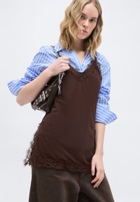 Νεαρό άτομο που φοράει ριγωτό μπλε πουκάμισο με στρώσεις καφέ φόρεμα με δαντέλα, κρατώντας μία σκούρα καφέ τσάντα με ασημένιο λουράκι αλυσίδα.