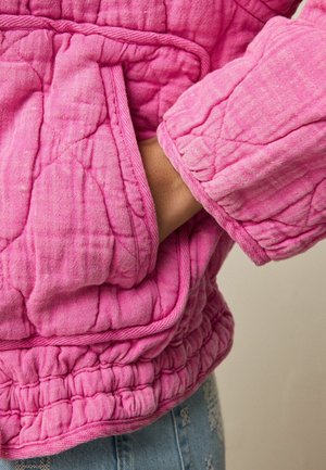 Hand in der Tasche einer gesteppte, leuchtend pinken Jacke mit gerafftem Saum und strukturierter Naht, getragen über blauen Denim.