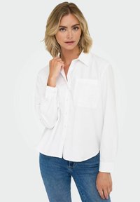 White button-up shirt with a chest pocket, featuring a subtle embroidered design, made from a smooth fabric, paired with blue jeans.