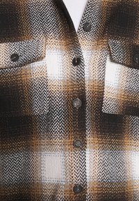 Hnědá, černá a bílá kostkovaná bunda s černými knoflíky a zapínacími náprsními kapsami na bílém vnitřním tričku.