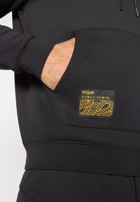 Black sweatshirt with a front pocket. Features a textured fabric, elastic cuffs and waistband, and a gold logo patch with a snake design.