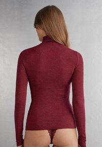 Burgundské tričko s dlouhým rukávem a vysokým límcem, vyrobené z texturované látky; má přiléhavý střih a minimální švy.