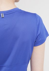 Osoba v modré košili s krátkým rukávem, která má texturovanou zadní část a malou černo-bílou pruhovanou cedulku poblíž výstřihu.