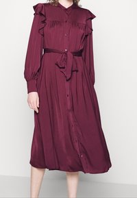 Dlouhá bordó šaty s volánovými rameny, páskem v pase, zapínáním na knoflíky a dlouhými rukávy vyrobené z hladké, splývavé látky.